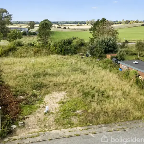 En tom byggegrund dækket med højt, vildt græs ligger ved siden af en beboelse og garage. Omgivelserne inkluderer åbne marker og nogle træer, hvilket giver et landligt præg.