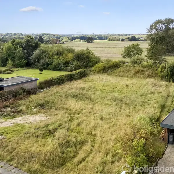 Græsklædt grund ligger stille mellem to huse i et landligt område med træer og marker i baggrunden. Grunden ser uplejet ud og grænser op til en vej.