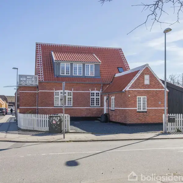 Et rødt murstenshus med rødt tag og hvide vinduer står på en hjørnegrund omgivet af et hvidt stakit, mens biler kører på vejen ved siden af.