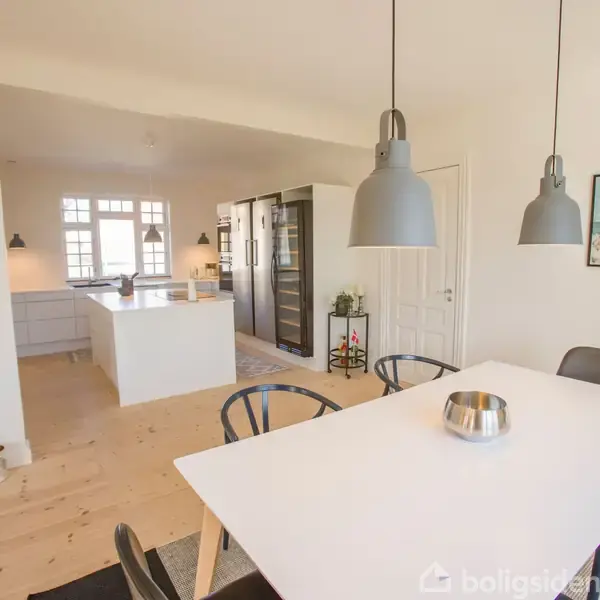 En hvid spisebord med skål står i et lyst køkken-alrum. Omkring er der stole, store lamper, en reol og et billede på væggen. Køkkenet har hvidt interiør med ø.