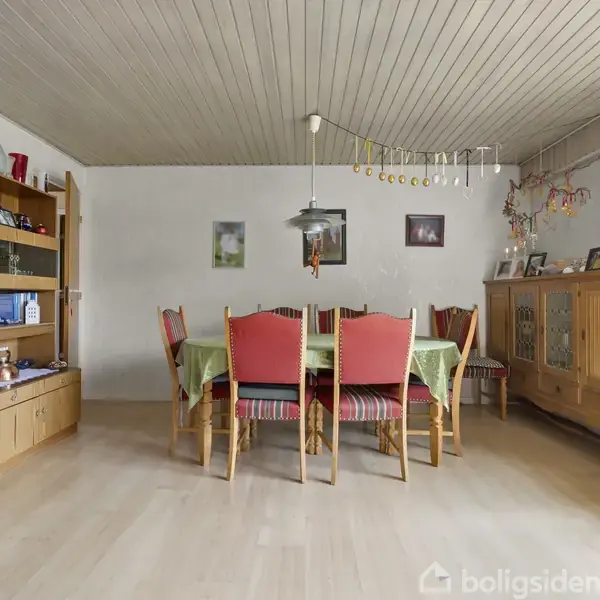 Spisestue med et spisebord dækket af en grøn dug omgivet af seks stole. Væghylder fyldt med bøger og glas til venstre; kommode med lamper og dekorationer til højre.