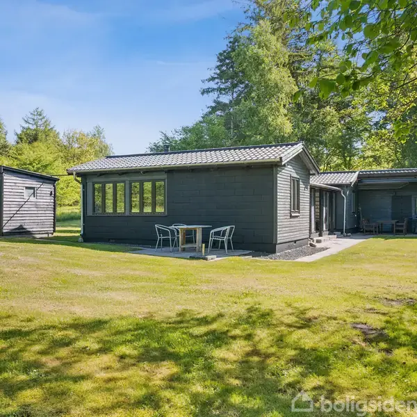 Et træsommerhus med gråt tag står på en grøn græsplæne omgivet af skov. Et lille skur ses til venstre, og der står havemøbler foran huset.