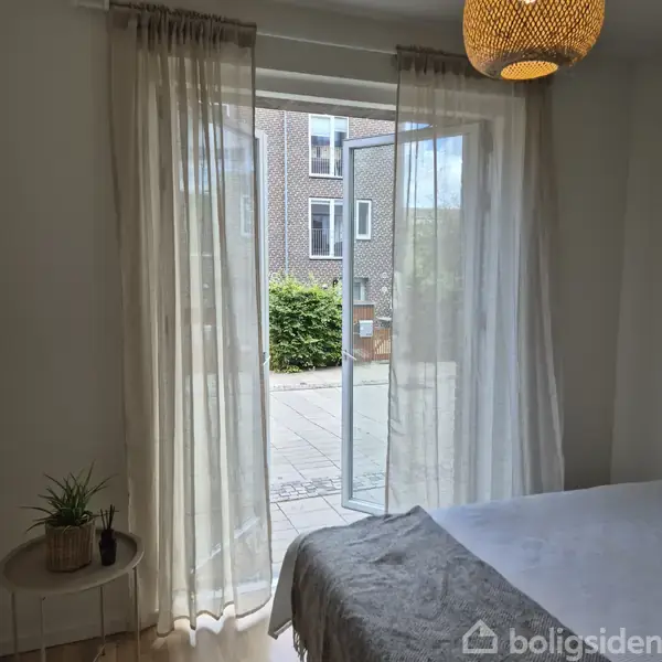 Et åbent vindue med hvide gardiner afslører en gård med flisebelægning og buske. Udenfor ses en murstensbygning. En seng og en plante er synlige indendørs.