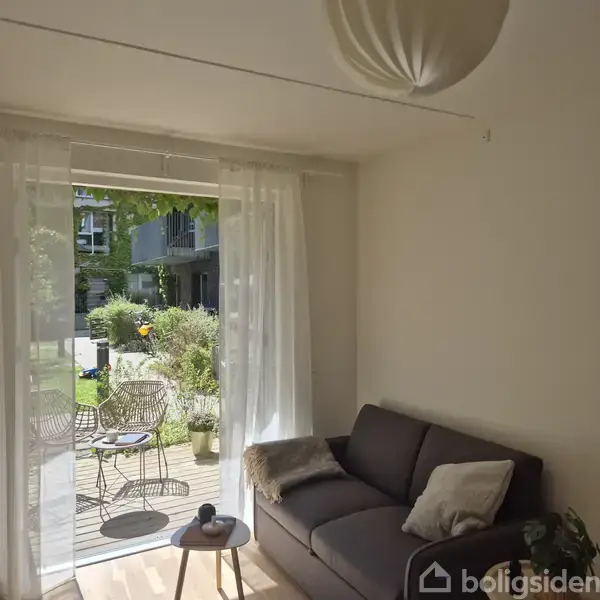 Sofa står inde i stue med udsigt til terrasse gennem åbne skydedøre med hvide gardiner. Terrasse har to stole og planter, omgivet af grønne buske og boligkompleks i baggrunden.