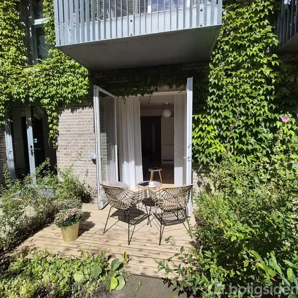 To stole står ved et lille rundt bord på en terrasse omgivet af frodige grønne planter. Terrassedørene er åbne ind til en bygning dækket af klatreplanter.