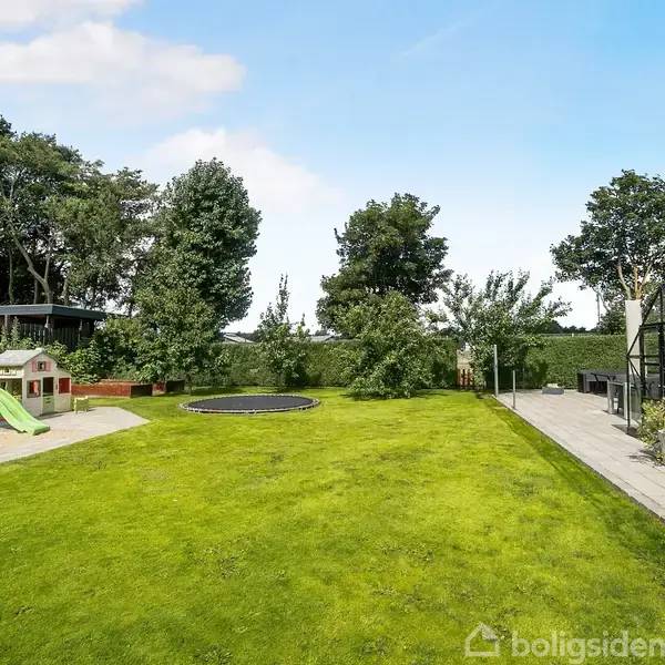 En legeplads med rutsjebane og sandkasse står i en have, hvor en trampolin er indlejret i græsplænen. Til højre ses en terrasse og et drivhus omringet af træer.