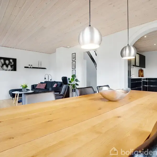 Et træbord står i forgrunden med en skål på, mens tre lamper hænger over det. I baggrunden er en stue med sofaer, planter og et åbent køkken.