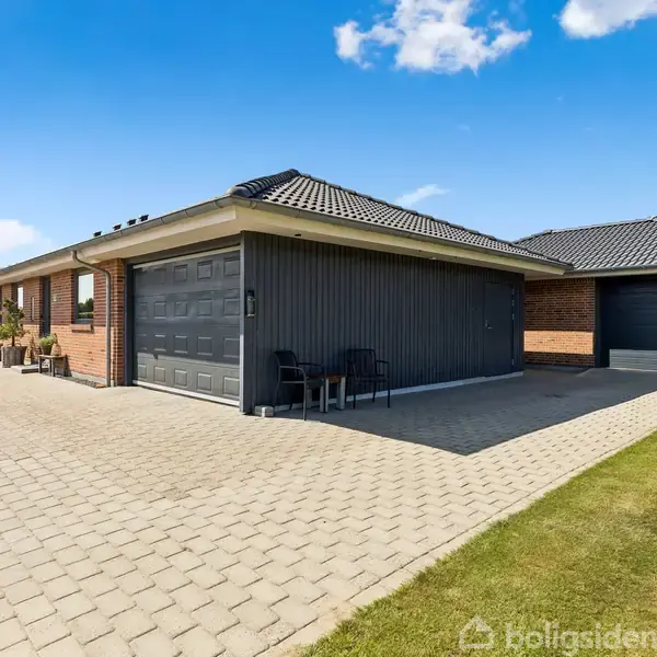 Et moderne etplanshus med integreret garage står på en flisebelagt indkørsel. Terrassemøbler er placeret ved siden af huset. Græsplænen omgiver hele området under en klar blå himmel.