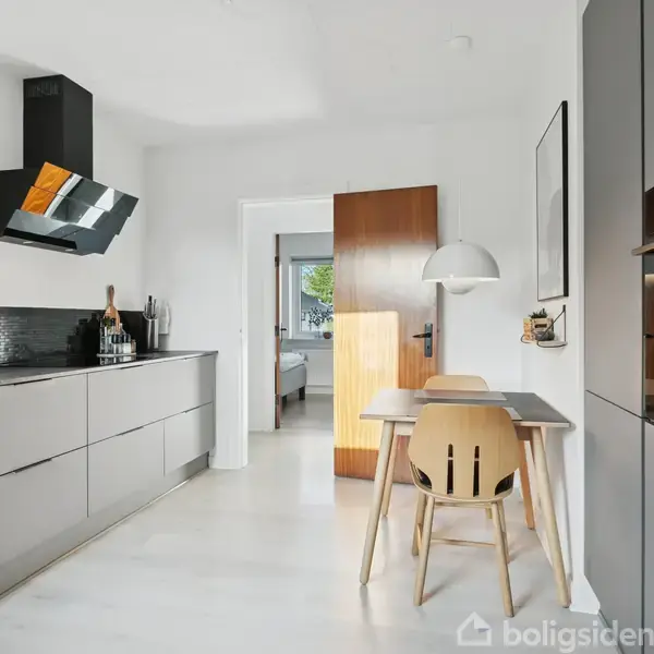 Et moderne køkken med grå kabinetter og indbygget ovn ved siden af et lille spisebord og stole. En åben dør fører til et lysfyldt rum i baggrunden.