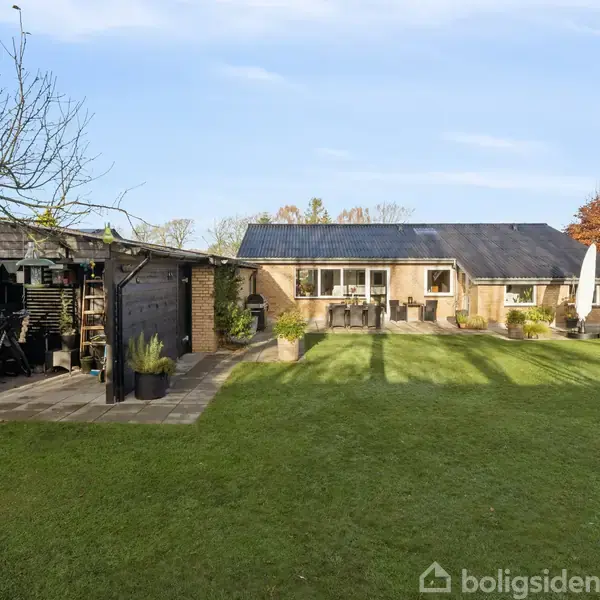 En enkeltstående bolig med terrasse og have omkranset af træer. Terrassen er delvist overdækket. Græsplænen er velplejet, og der er placeret havemøbler nær huset og en parasol.