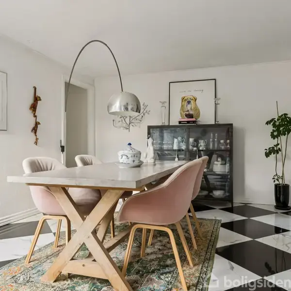 Et spisebord med lyserøde stole står i et rum med sort-hvide ternede gulve. Et stort lampehoved svæver over bordet. Vægge er dekoreret med kunst og planter.