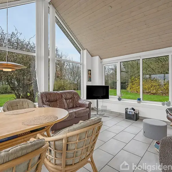 Stue med rattanmøbler og sofahjørne, placeret nær store vinduer. Udsigten viser en grøn have med buske og træer, mens hvide fliser og et brunt loft dominerer interiøret.