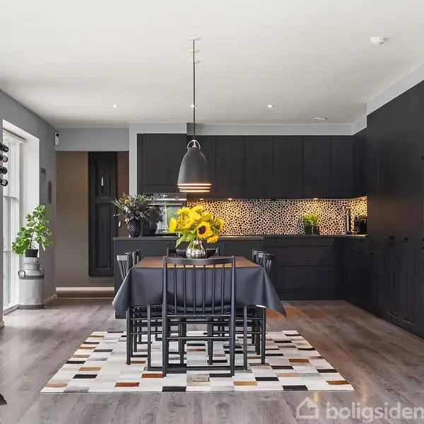 Et spisebord med sorte stole står midt i et køkkenalrum. Bordet er dækket med en mørk dug og solblomster i en vase. Køkkenet har sorte skabe og mønstret vægbeklædning.