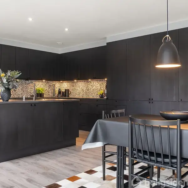 Et moderne køkken med sorte skabe og øer, dekoreret med mosaikfliser på væggen. En sort spisebord står ved siden af med sorte stole. Klare lamper hænger over bordet.