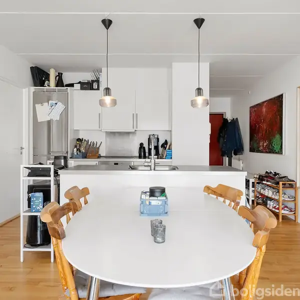 Et spisebord med træstole står i forgrunden. Køkkenet i baggrunden har hvide skabe. To loftlamper hænger over bordet. Der er en guitar på væggen til højre.