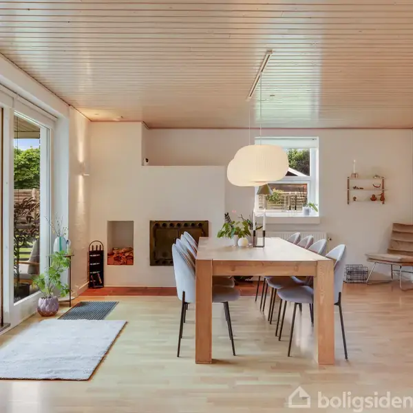 Stor, lys spisestue med træspisebord og stole. En moderne lampe hænger over bordet. Rummet har trægulv, vinduerne åbner til en have. Vægge er hvide med enkelte dekorationer.