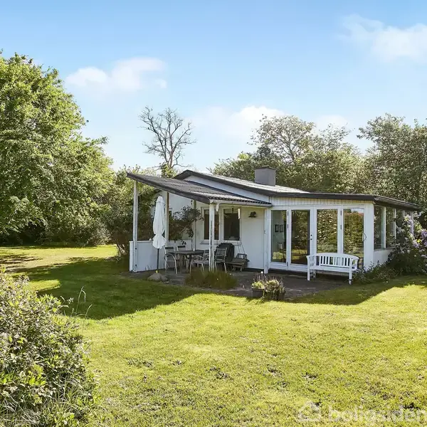 En hvid villa med en overdækket terrasse og havemøbler står i en frodig, grøn have omgivet af træer under en blå himmel.