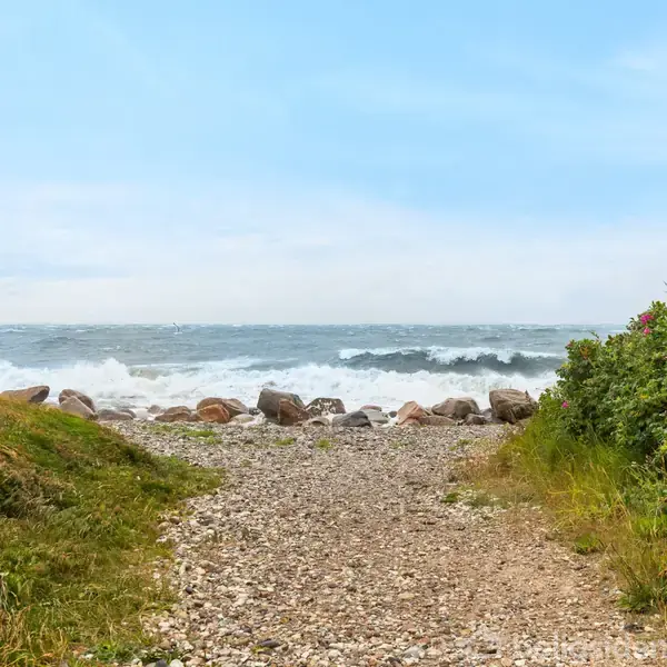 En stenstrand med bølger, der slår mod kysten. Omgivet af grønne buske og græs, under en klar blå himmel.