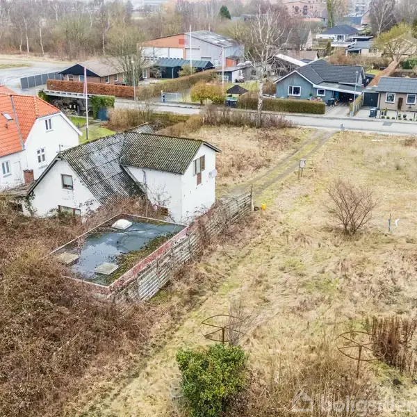 Et gammelt, hvidt hus med grønt tag står forladt. Omkring det er der en stor, tilgroet grund i en villakvarter. Flere huse ses i baggrunden.