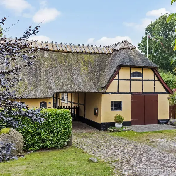 Bindingsværkshus med stråtag står i en grøn have omgivet af træer og buske. En grusvej og brosten leder til en garage med store, brune døre.