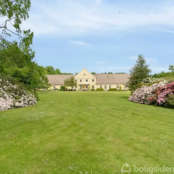 En stor, gul bygning står i baggrunden, omgivet af en stor, frodig græsplæne. Farverige buske blomstrer på begge sider, mens skovelementer omgrænser området.