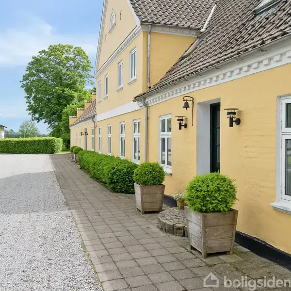 Gul bygning med hvidrammede vinduer står stille ved siden af en grusvej. Foran døren er der potteplanter, og i baggrunden ses grønne træer og buske.