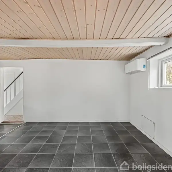 Et tomt rum har sorte fliser på gulvet, en træloft og en hvidmalet væg. En lille vinduesåbning viser et udendørs landskab, og der er en dør til en trappe.