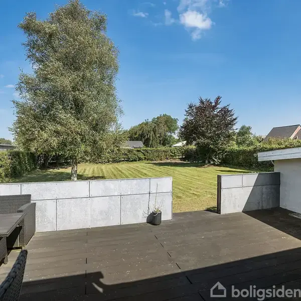 Terrasse med bord og bænke står omgivet af en have med græs og træer. I baggrunden ses huse og en mast under en blå himmel.