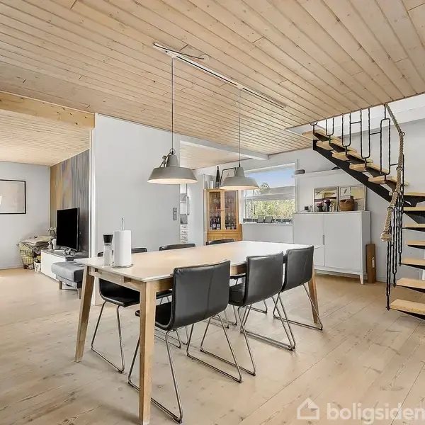 Spisebord med stole står i et åbent rum, oplyst af loftlamper. Til højre er der en trappe, og i baggrunden ses en stue med sofa og tv.