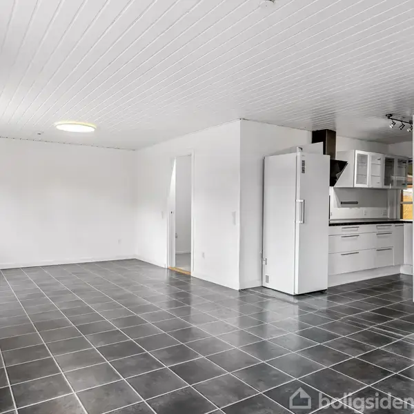 Et lyst, tomt rum med sorte fliser, en hvid væg og loft. Køkkenet har hvide skabe og en køleskab i hjørnet. Dør fører til et tilstødende rum.