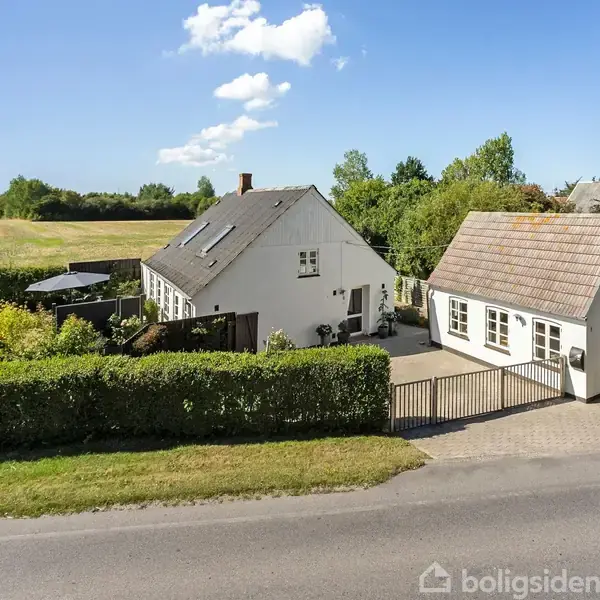 Et hvidt hus og en garage står ved vejen, omgivet af hække og grønne træer. Sollys belyser området, og en klar blå himmel lægger sig som baggrund.