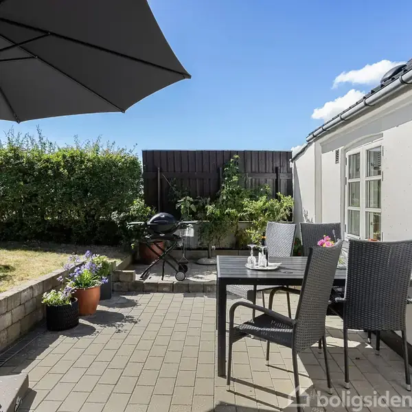 En terrasse med en sort parasol skygger over et bord og stole. En kuglegrill står ved siden af, omgivet af blomsterkrukker. Baggrunden har grønne buske og en hvid bygning.