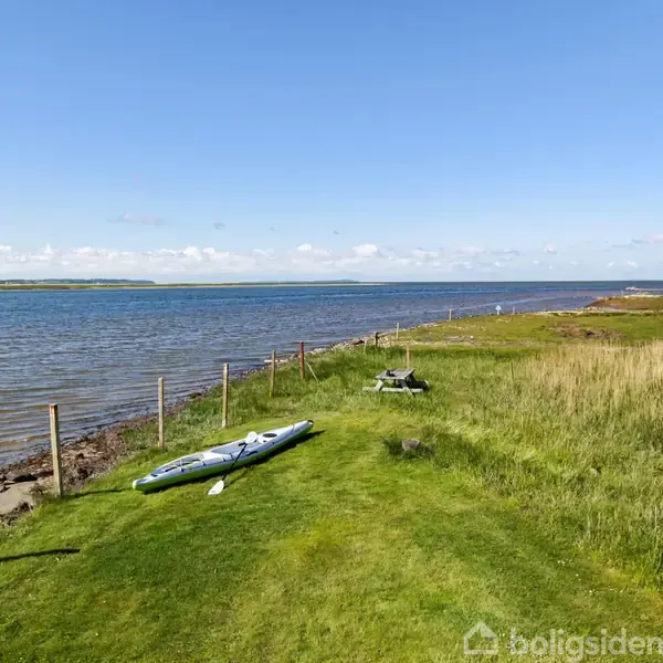 En oppustelig kajak ligger på græsplænen ved kysten. Bagved ses et hegn langs vandet med en klar blå himmel og spredte skyer i baggrunden.