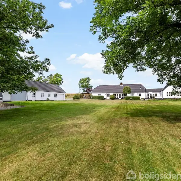 Grøn græsplæne strækker sig mod hvid landejendom med sorte tage. Omkringliggende træer kaster skygger på plænen. Blå himmel med få skyer ses i baggrunden.