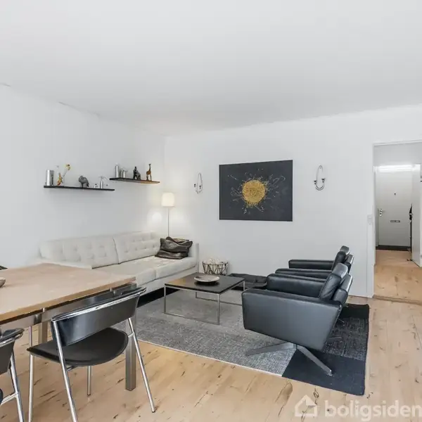 Stue med en hvid sofa og to sorte lænestole omkring et sofabord på et tæppe. En spisebord med stole står til venstre. Væggene har moderne kunst.