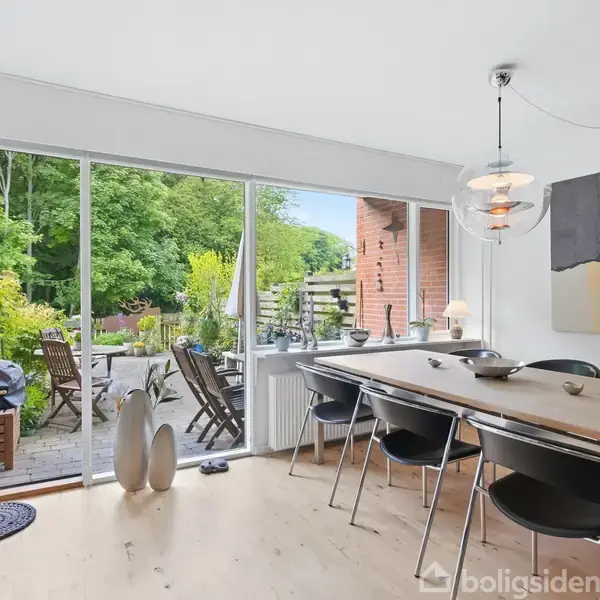 Spisebord med sorte stole står i et lyst rum med udsigt til terrasse gennem glasdøre. Udenfor ses havemøbler og grøn have. Moderne kunst hænger på væggen.