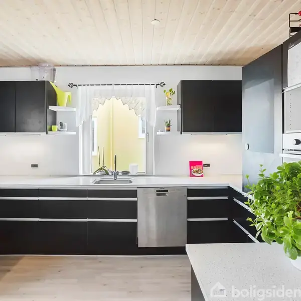 Sort køkken med L-formet bordplade, hvide skabe, en vask i midten og grønt planteelement. Ved siden af vasken står en opvaskemaskine. Vinduet har en dekorativ gardinkant.