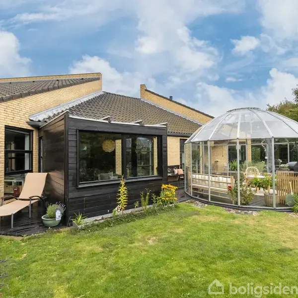 Et hus med gule murstensvægge har en tilstødende glaskuppelstruktur. En træterrasse med liggestol og planter omringes af græsplæne og buske under en blå himmel med skyer.