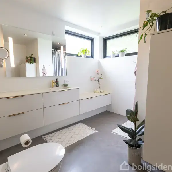Et moderne badeværelse med hvide skabe og spejl langs væggen. Planter dekorerer rummet, og naturligt lys kommer ind gennem to store vinduer. Toilet og tæpper er synlige på gulvet.