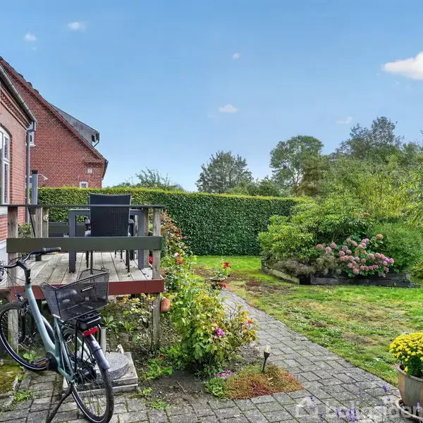 Cykel står parkeret ved siden af en terrasse med bord og stole. I baggrunden ses en velplejet have omgivet af buske og træer under en klar, blå himmel.