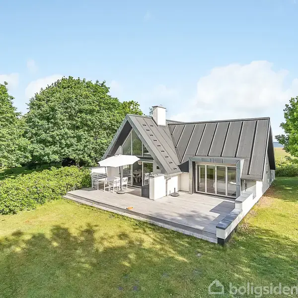 Hus med moderne tag, står på en terrasse med parasol og havemøbler. Omgivet af grønne træer og græsplæne under en blå himmel.