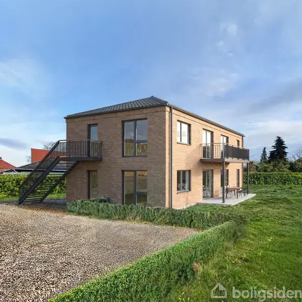 Et moderne, to-etagers hus med murstensfacade har en udvendig trappe og flere altaner. Det står i en grøn have med grusplads forud, under en skyfri blå himmel.