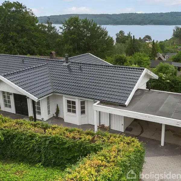 En hvid villa med mørkt tag står stille omkranset af grønne buske, med en carport til højre. Baggrunden viser en sø med træer i en bakkelandsby.