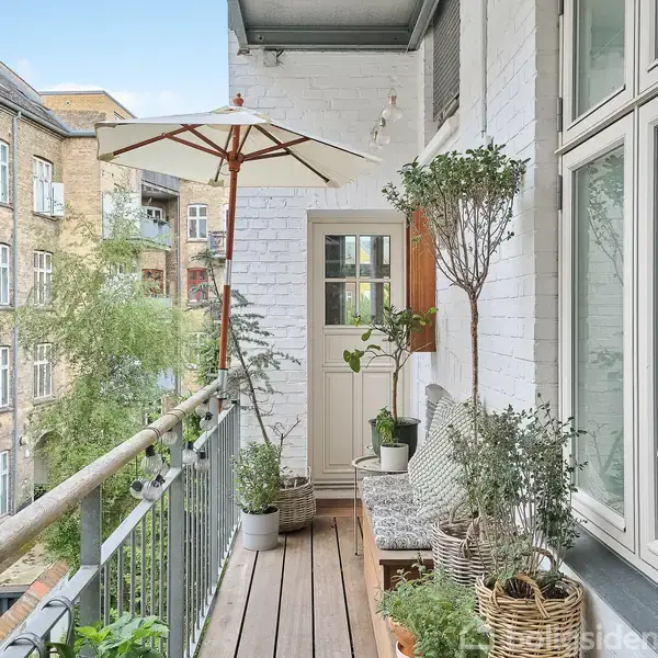 Balkon med træplanker og planter i potter, beliggende udendørs. En parasol giver skygge, og en hvid dør fører indenfor. Omgivet af murstensbygninger i et byområde.
