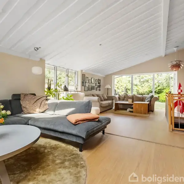 Sofa står i en lys stue med skrå loft. Den er placeret ved siden af et rundt bord med blomster. Store vinduer giver udsigt til en grøn have.
