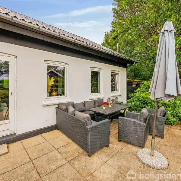 Terrassemøbler står på en flisebelagt terrasse ved siden af et hvidt hus. Møblerne omfatter en sofa, stole og et bord med blomster på. En parasol er slået op ved siden af.