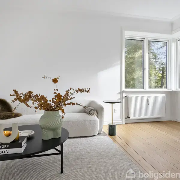 Sofa med puder står ved siden af vinduer, der lukker lys ind i stuen. Bord med vase og bøgerne "New York" og "London" foran sofaen.