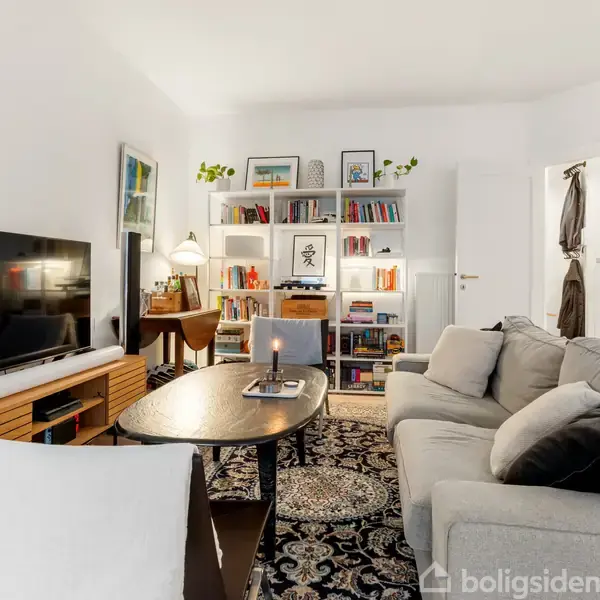 Stue med en grå sofa foran et rektangulært sofabord med en tændt candle. Et stort fjernsyn står til venstre. Reoler med bøger og pynt i baggrunden. Lys og planter skaber en hyggelig atmosfære.