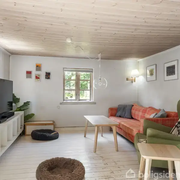 Stue med en orange sofa og et grønt sofabord foran, placeret på et lyst trægulv. Et TV står til venstre på et hvidt møbel. Vægge dekoreret med billeder.