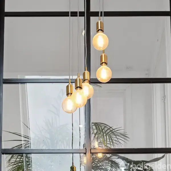 Fire hængende pærer lyser i en række, monteret i gyldne fatninger. De hænger foran et vindue med en indendørs plante delvist synlig i baggrunden.